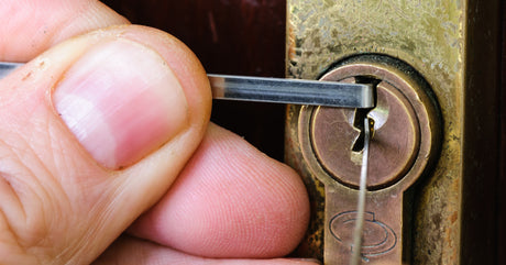 Closeup of someone’s fingers holding lock-picking tools as they pick an older-looking bronze lock on a door.