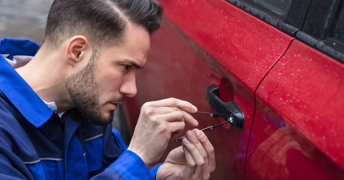 Close-up of a male automotive locksmith using two lock-picking tools to unlock the door of a red vehicle.