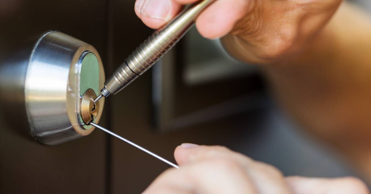 A closeup of someone picking the lock on a door. They are using a pick and a tension wrench to open the lock.