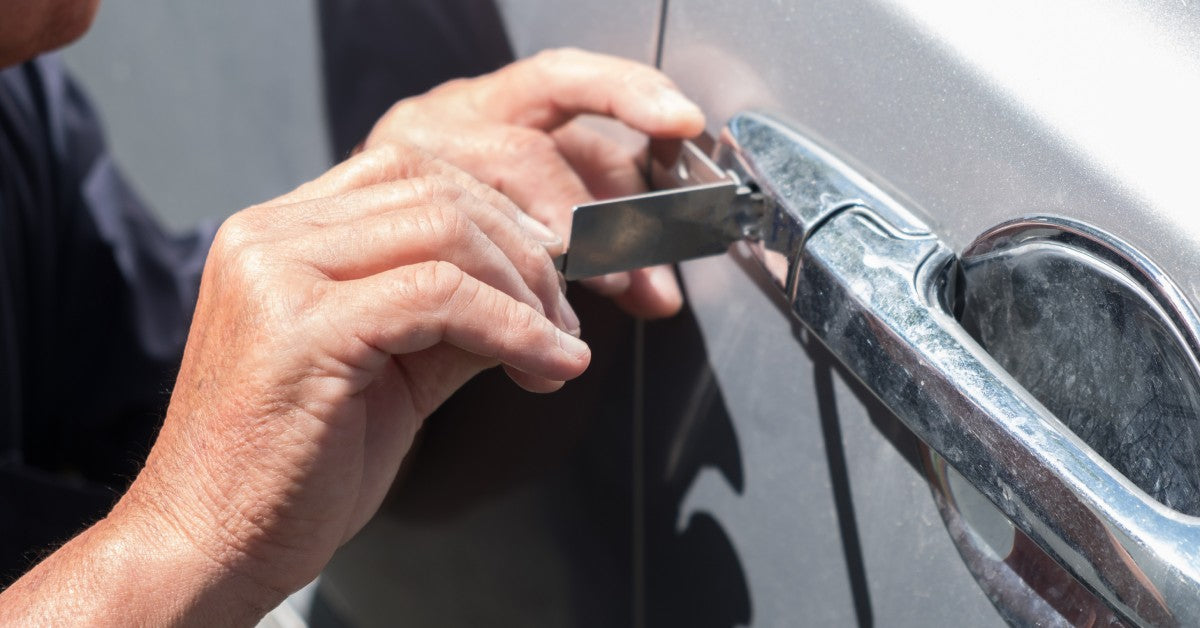 A close-up of a man picking the lock on a silver car with a lock pick. The metal door handle is covered in blemishes.