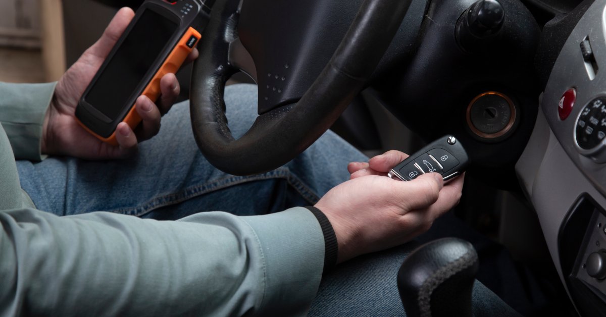 A man sitting in the driver's seat of a car with a smart key in one hand and a programming device in the other.